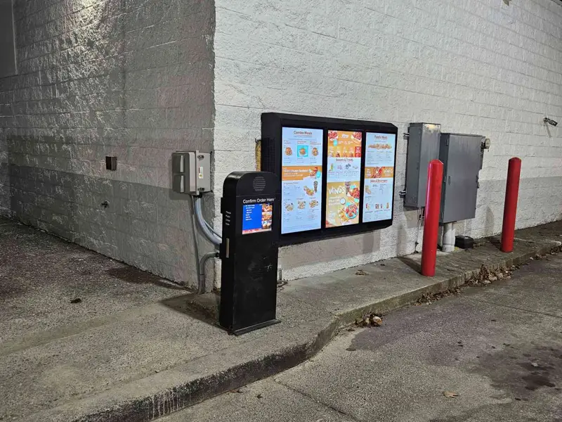 Wall-mounted three-panel drive-thru board and confirm-order pedestal lit at night