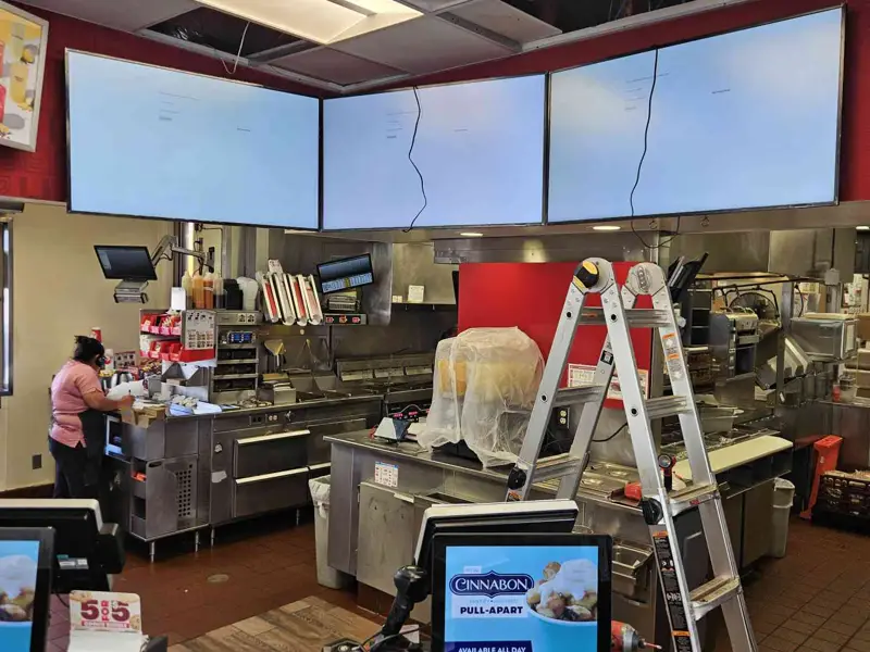 Three overhead screens powered above a fast-food counter, ceiling tiles open and a ladder in place
